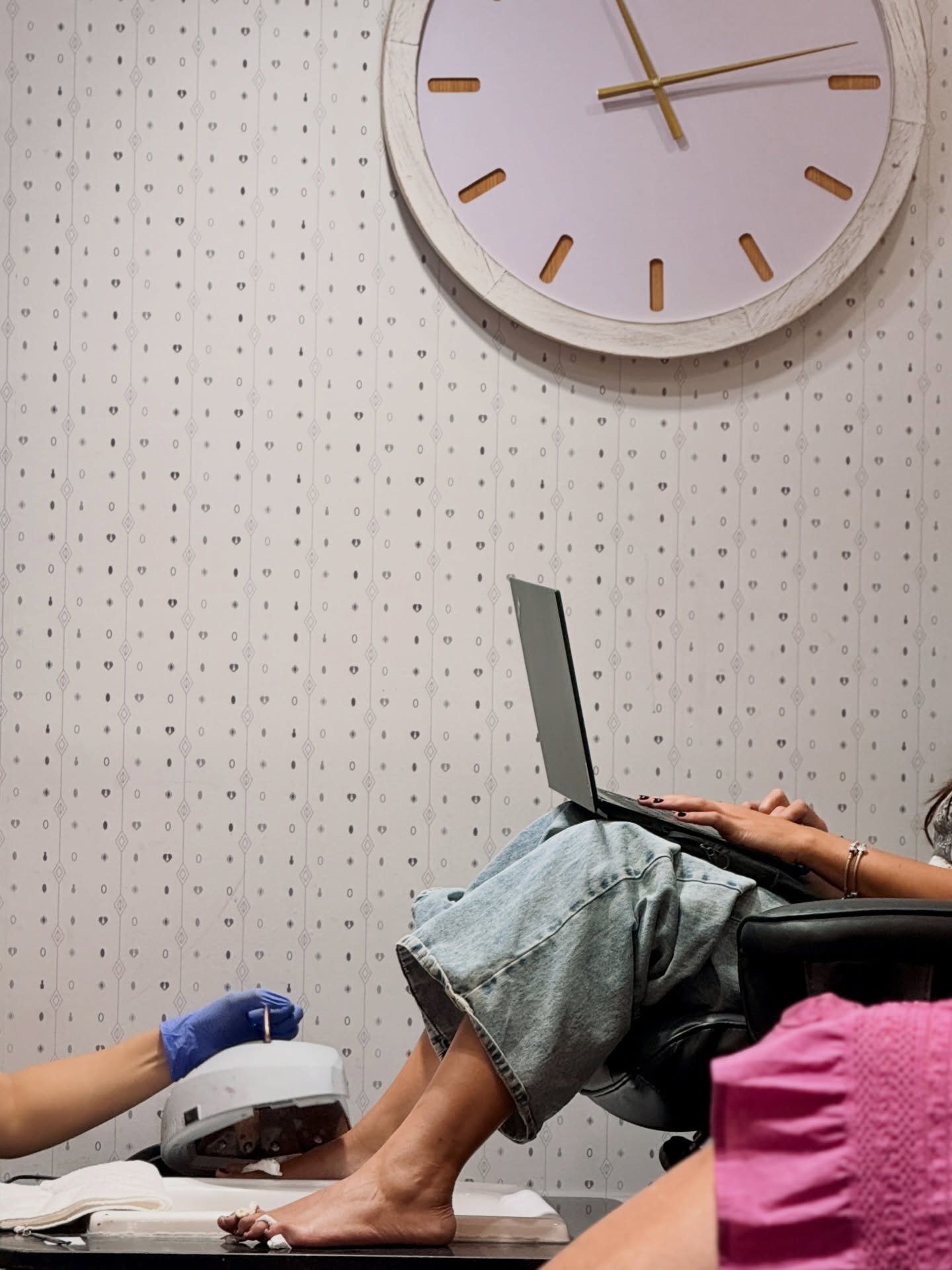 Una clienta trabaja con su notebook en el sillón de pedicura mientras le secan el esmaltado, bajo el gran reloj de pared del salón